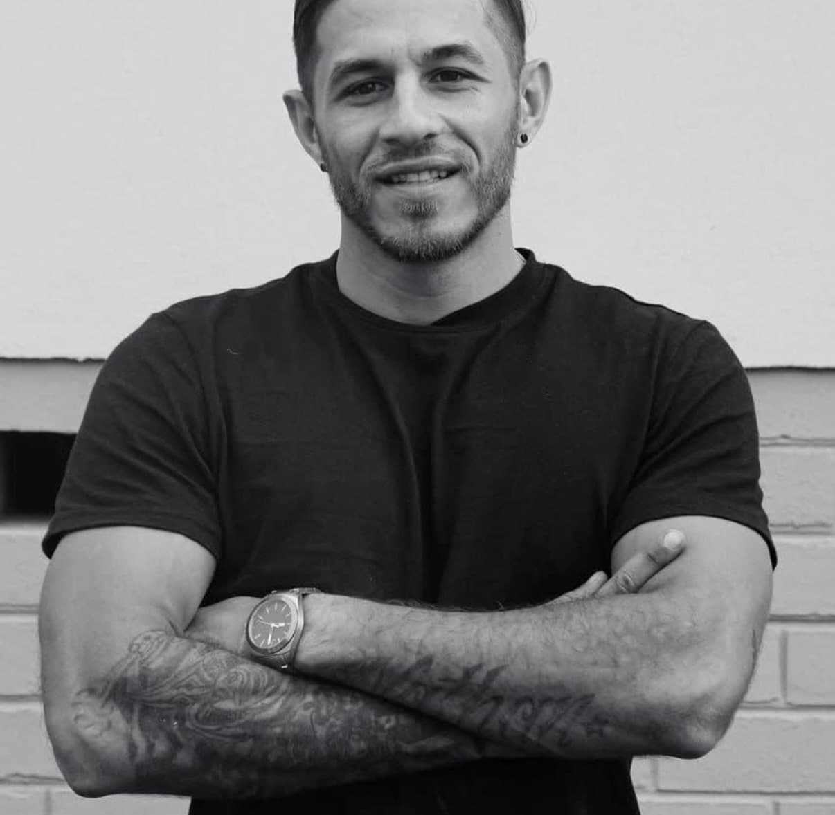 Smiling man with a beard and tattooed arms crossed in a black and white portrait.